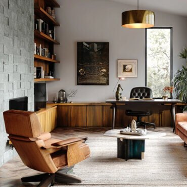 Mid-century modern home office and living room featuring leather lounge chair, peach sofa, wooden shelves, and large windows with black frames.