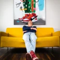 Huge colorful hip hop art of digitally drawn Air Jordans on a person wearing green and white checkered pants hangs above yellow couch. On the couch, a man casually leans back, chin tilted towards the sky. On his feet are red Air Jordans, which are similar to the sneakers drawn in the wall art.