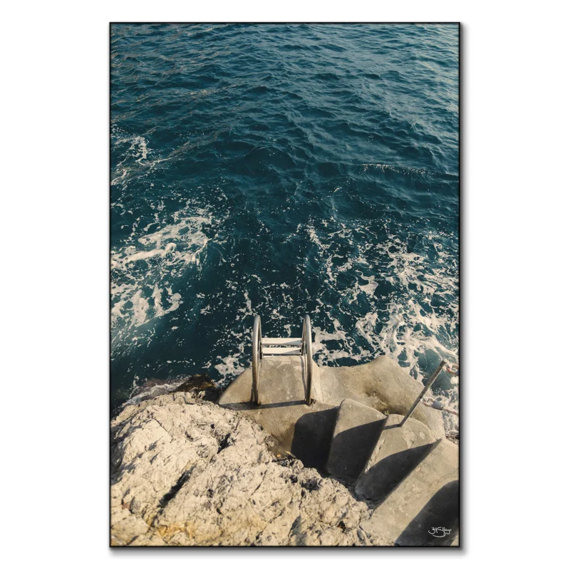 A coastal photograph featuring a stone staircase leading to a metal ladder descending into the deep blue sea with foamy waves.